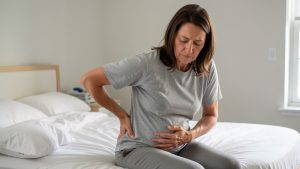 Femme adulte assise sur le bord d’un lit, une main posée dans le bas du dos et l’autre sur l’abdomen, illustrant des douleurs associées à la constipation.