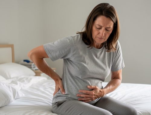 Femme adulte assise sur le bord d’un lit, une main posée dans le bas du dos et l’autre sur l’abdomen, illustrant des douleurs associées à la constipation.