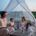 Une famille en vêtements longs clairs profite d’une terrasse dans les Pays de la Loire au crépuscule, avec une moustiquaire et un répulsif posés sur la table.